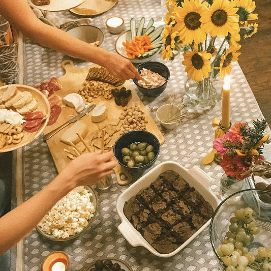 A colorful table of snacks and small bites with sunflowers, lit candles, and hands reaching to fill their plates.