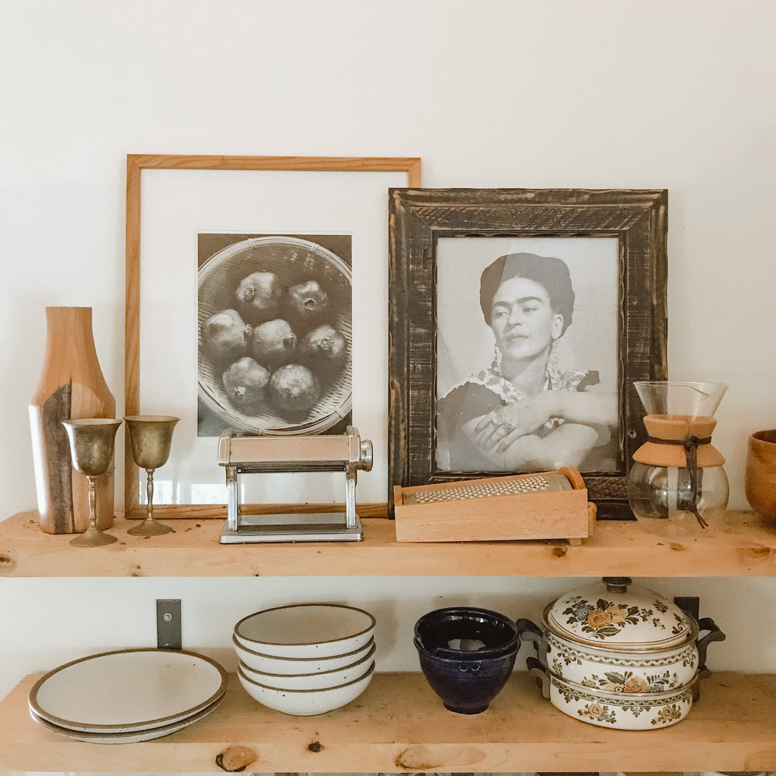 Kitchen shelves containing framed black and white photos of pomegranates and Frida Kahlo, a pasta machine, boxed cheese grater and various dishes