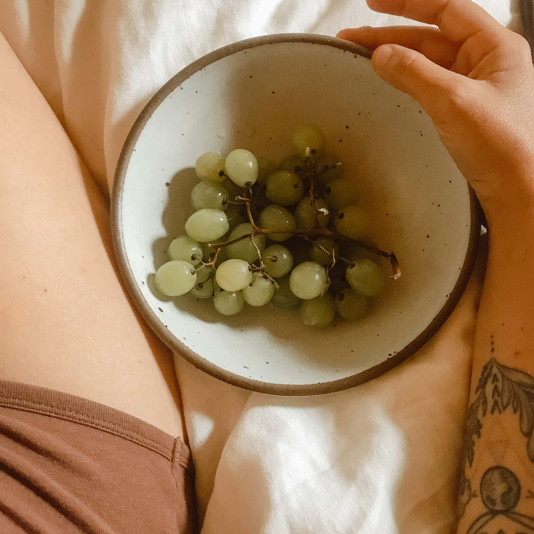 An above view of a leg and an arm laying on a white bed with a bowl of green grapes