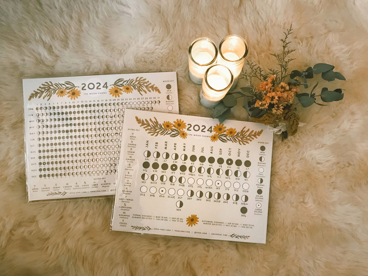 Hina Luna's 2024 moon calendar designs laying on top of a white sheepskin rug surrounded by candles and a dried flower bouquet